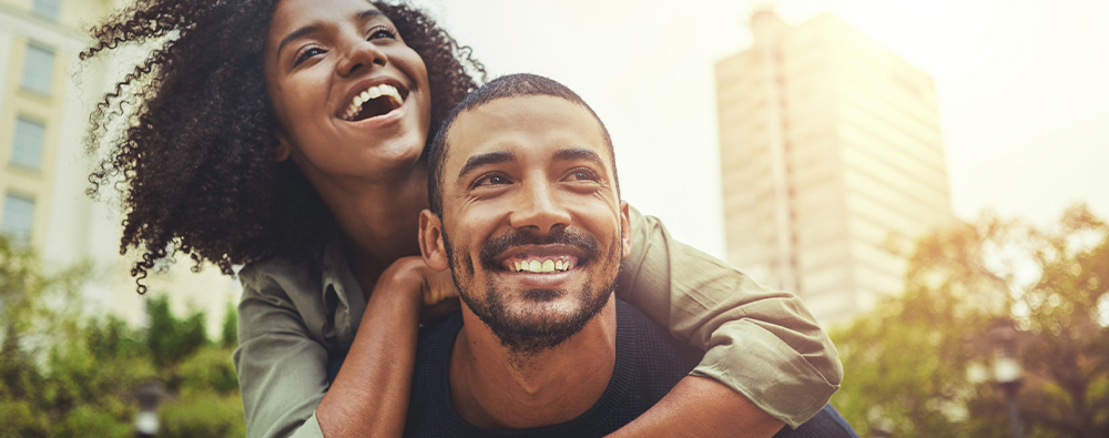 An African American woman riding piggyback on her boyfriend in the downtown area of a city and both are smiling.