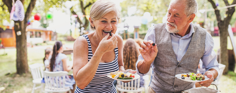 An older couple smiles as they grab a plate of food at a picnic.