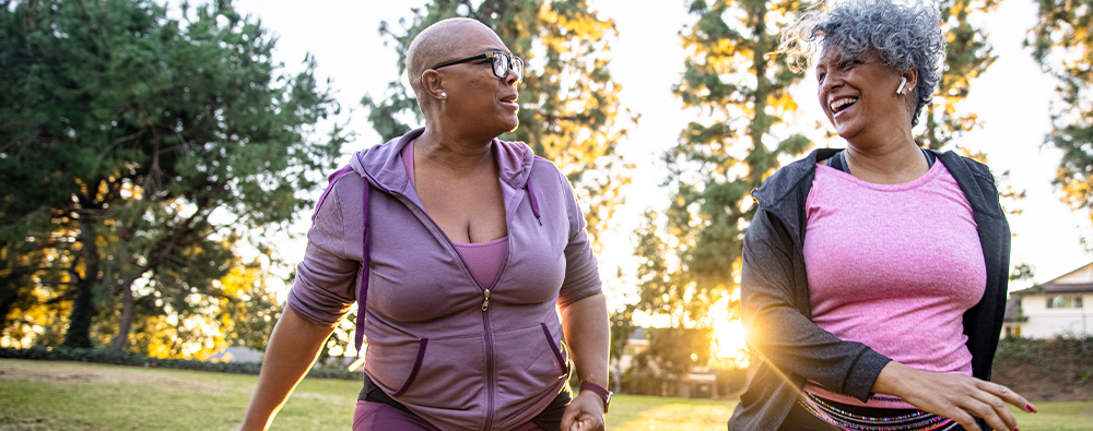 Two older black women walking in the park on a sunny day, both are smiling and talking. 