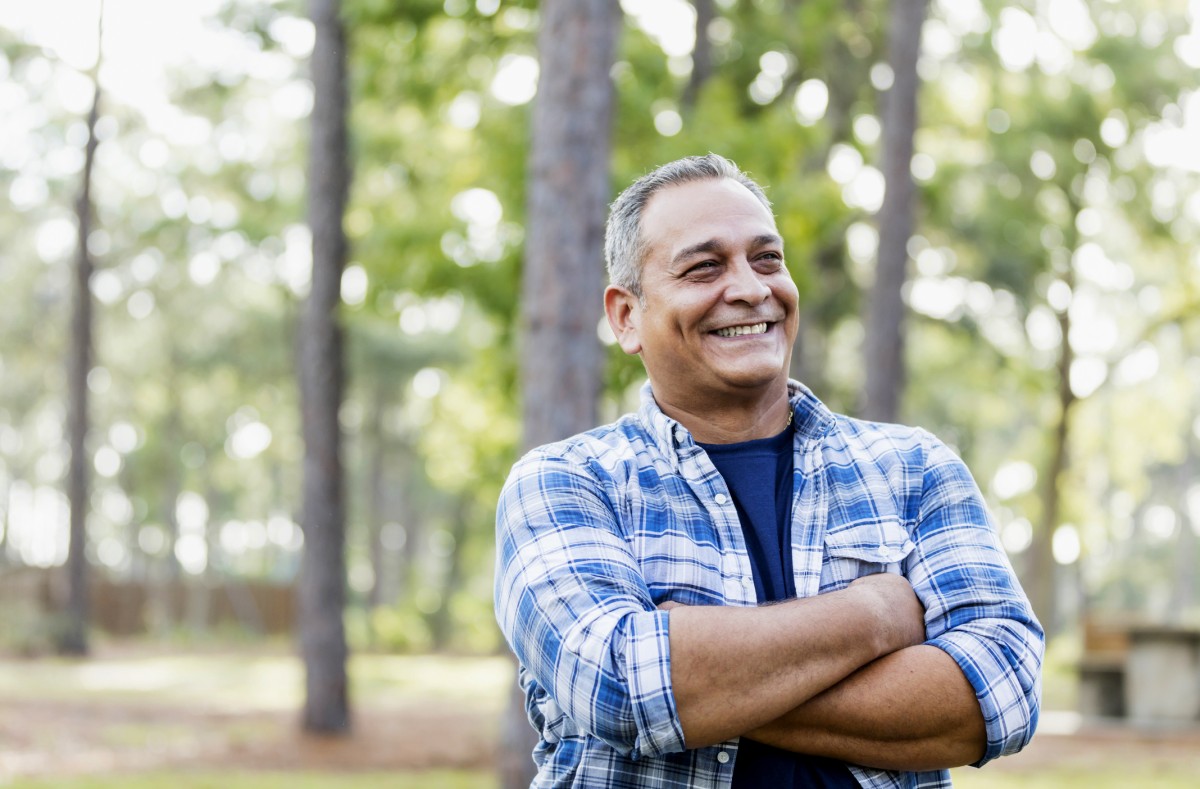 Man smiling with his arms folded in a wooded setting
